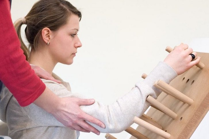 Eine Patientin uebt mit Unterstuetzung einer hinter ihr stehenden Therapeutin Greifen an einem schraegen Brett mit Holzstaeben in der Handtherapie im VAMED Rehazentrum Karlsruhe