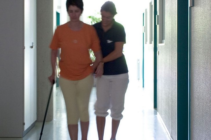 Eine Patientin laeuft mit zwei Gehhilfen und Unterstuetzung einer Physiotherapeutin einen Gang entlang im Rahmen der Gangschule nach Bobath in der Physiotherapie Karlsruhe