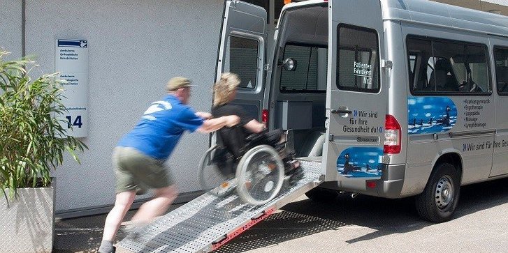 Fahrdienstmitarbeiter schiebt Patient im Rollstuhl über eine Laderampe ins Auto