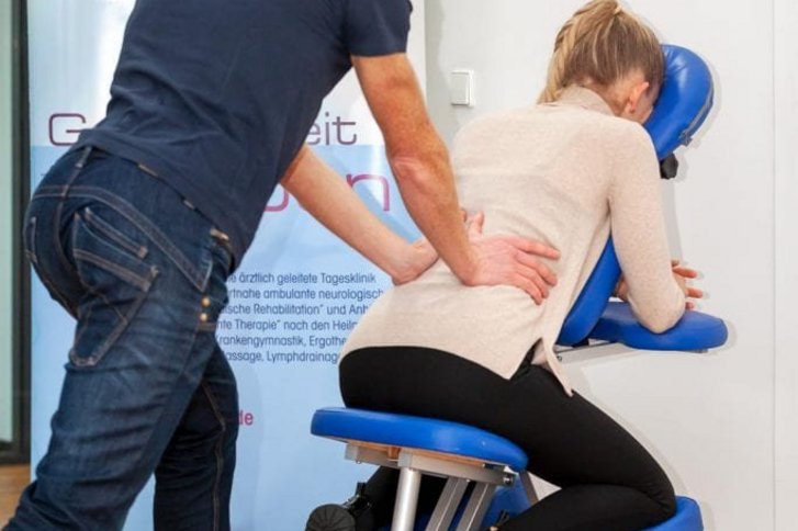 Eine nach vorne aufgestuetzte Patientin erhaelt eine Massage am unteren Ruecken durch einen Physiotherapeuten