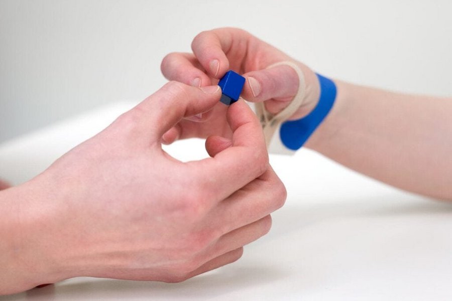 Nahaufnahme einer geschienten Patientenhand die im Pinzettgriff eine kleine Kugel aus den Fingern eines Therapeuten nimmt