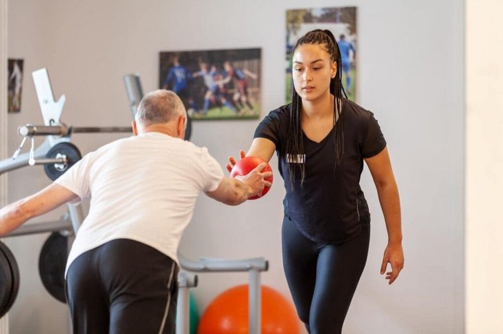 Blick auf eine Trainingseinheit im Sport- und Gymnastikkurs bei der eine Therapeutin in Frontalansicht einen Ball an einen von hinten gezeigten Patienten im Ausfallschritt übergibt